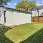 A fenced backyard with a small light-gray shed, green grass, a metal climbing dome on the left, and a few trees and houses in the background under a blue sky.