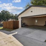 A cream-colored detached garage with an open door, revealing bicycles, shelves, and boxes inside. A wooden fence borders the property, and trees and houses line the street in the background.