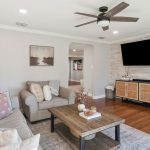 A cozy living room with a gray sofa, armchair, and a wooden coffee table. A TV is mounted on a light tile accent wall above a wood console. There are plants, decorative items, and neutral-toned decor throughout the space.