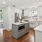Modern kitchen with white cabinets, stainless steel appliances, a gray island, built-in microwave, farmhouse sink, and wood flooring. Bright natural light enters from large windows and an adjacent open living area.