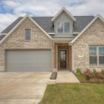 A single-story brick house with a triple garage, a front porch, large windows, a manicured lawn, and flower beds, set under a partly cloudy sky.