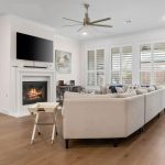Modern living room with light walls and wood floors, featuring a beige sectional sofa, a fireplace with a TV above, wall art, a ceiling fan, a small table, and large windows with white shutters.