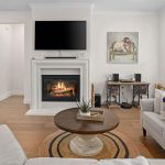 A cozy living room with a lit fireplace, a TV above it, and a bull painting on the wall. There's a round coffee table, sofas with blue-and-white pillows, and large windows with white shutters.