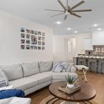A modern open-concept living room and kitchen with a light gray sectional sofa, decorative pillows, a round coffee table with a plant, wall art, wooden floors, and a breakfast bar with three stools.