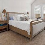 A cozy bedroom with a wooden bed, white bedding, and multiple pillows. A nightstand with lamps, books, and a colorful bird painting sits beside the bed. An exercise bike is near large windows with white shutters.