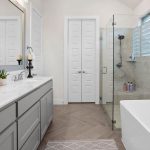 Modern bathroom with light gray cabinets, dual sinks, a glass-enclosed shower, white freestanding bathtub, and decorative accents on the countertop. The room features neutral colors and natural light from a window.