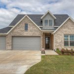 A modern two-story brick house with three-car garage, gray doors, a small front porch, manicured lawn, and flower beds under a partly cloudy sky.