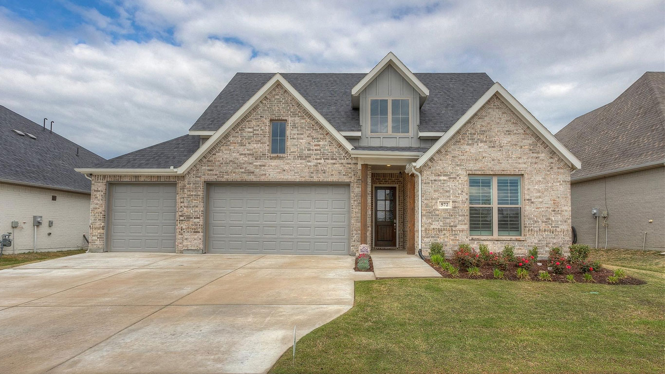 A modern two-story brick house with three-car garage, gray doors, a small front porch, manicured lawn, and flower beds under a partly cloudy sky.