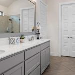 Modern bathroom with light gray cabinets, double sinks, marble countertop, large wall mirror, and glass-enclosed shower. Decorative items and candles sit on the counter. Light wood herringbone floor and white walls.
