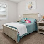 A tidy bedroom with a light wooden bed, blue bedding, stuffed toys, two bedside tables with lamps, a teal desk and chair, a window with white shutters, and artwork above the bed.