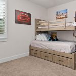 A tidy bedroom with a wooden bunk bed and light gray bedding, drawers under the bed, a desk with a computer, wall art including a red car photo, and a window with white shutters.