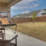 Two wicker chairs sit on a covered patio overlooking a backyard. A basketball arcade game is next to the chairs, and the yard is enclosed by a wooden fence with a few houses visible in the background.
