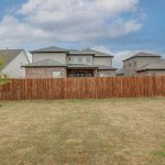A fenced backyard with dry grass, a small tree on the left, a trampoline on the right, and several houses in the background under a partly cloudy sky.