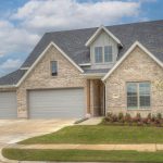 A modern, two-story brick house with a large driveway, three-car garage, front lawn, flower bed, young tree, and a cloudy blue sky in the background.