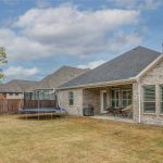 A backyard with dry grass, a trampoline, a covered patio with lounge chairs, a grill, and a brick house with shuttered windows under a partly cloudy sky.