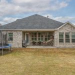 A backyard with a brick house, covered patio, grill, chairs, hanging chair, and a trampoline on the left, with a wooden fence and neighboring houses visible.