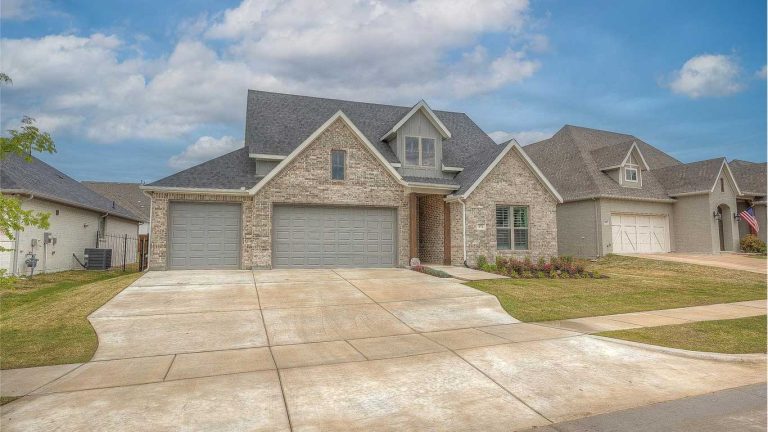 A modern two-story brick house with a three-car garage, sloped roof, and neatly landscaped front yard under a partly cloudy sky in a suburban neighborhood.