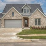 A modern two-story brick house with three gray garage doors, a small covered porch, front lawn, flower beds, and a concrete driveway under a partly cloudy sky.