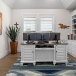 A home office with a white desk holding three monitors, a patterned rug, built-in shelves with books and decorations, a large potted plant, and western-themed artwork on the wall. Natural light enters through two windows.