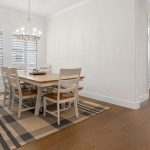 A bright dining room with a wooden table, six white chairs, a plaid area rug, a modern chandelier, and a sideboard. Large windows with shutters let in natural light; a hallway leads to a dark wood front door.