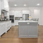 Modern kitchen with white cabinets, stainless steel appliances, gray island with white countertop, light wood flooring, and dining and living area with a fireplace and TV in the background.