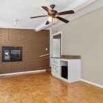 A narrow room with parquet flooring, a brick accent wall featuring a built-in fireplace, a ceiling fan, large windows, and a small white built-in cabinet with a granite countertop.