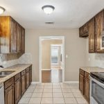 A modern kitchen with wooden cabinets, granite countertops, stainless steel appliances, a window above the sink, and a tiled floor. The room opens into a dining area with wooden flooring.