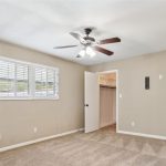 A carpeted, empty bedroom with beige walls, a ceiling fan with lights, two windows with white blinds, an open door leading to a walk-in closet, and ample natural light.