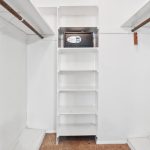 A white walk-in closet with wooden rods on both sides, white shelving in the center, and a black safe placed on the top shelf. The floor is carpeted.