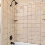 A tiled bathtub and shower area with two corner shelves, bronze fixtures, and a showerhead. The door to the bathroom is partially open, revealing white walls and dark handles.