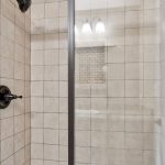 A tiled walk-in shower with a glass door, dark metal fixtures, and a built-in recessed shelf. White tiles cover the walls, and a three-bulb light fixture is visible outside the shower.