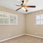 A small, empty room with beige walls, tan carpet, a ceiling fan with lights, and two windows with white shutters. The upper wall has a row of small white triangle decals.