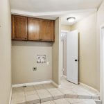 A small laundry room with beige walls, tile floor, wooden cabinets mounted above laundry hookups, a white door slightly open, and a thermostat on the wall.
