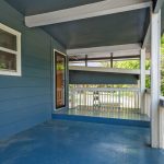 A covered porch with blue painted flooring and walls, white railing, a window, and a glass door, overlooking a green, leafy yard.
