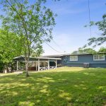 A backyard with green grass, a large tree, and a blue house with white trim. There is a covered patio area with a white railing and the yard is surrounded by lush green trees. The sky is clear and blue.