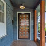 A modern front porch with blue brick walls, a wooden door featuring square glass panels, a tiled floor, a ceiling light, and wooden support beams, overlooking green bushes and trees.