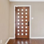 Modern wooden front door with a grid of square glass windows, set in a beige-walled entryway with dark wood floor; part of a wooden piano is visible on the right side of the image.