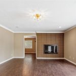 A spacious living room with beige walls, dark wood flooring, a brick fireplace, large windows on the right, and a chandelier on the ceiling. An open doorway leads to another room in the background.