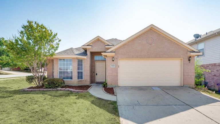 A single-story brick house with a two-car garage, large front windows, a light-colored front door, a small porch, and minimal landscaping with a tree and some bushes in the front yard.