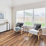 A bright, minimalist living room with two wooden armchairs, gray cushions, a side table, and a console table against the wall. Large window, wood flooring, and simple decor create an airy, modern feel.