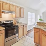Modern kitchen with light wood cabinets, granite countertops, stainless steel oven, electric stove, double sink, and large windows letting in natural light. There are green apples on the counter and a plant near the sink.