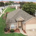 Aerial view of a single-story brick house with a two-car garage, neatly manicured lawn, and a curved walkway leading to the front door, located on a suburban street lined with trees and neighboring homes.
