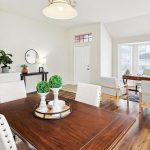 Bright, open living and dining area with wood floors, white walls, a wooden dining table with chairs, a small office nook by large windows, plants, and decorative accents throughout.