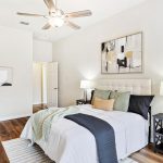 A modern bedroom with a bed featuring white and dark gray bedding, two bedside tables with lamps, abstract art above the bed, wood flooring, a ceiling fan, and minimal decor.