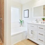 Bright bathroom with a glassless shower, tan tiles, a white bathtub, a double-sink white vanity, large mirror, and two small potted plants. Natural light comes through a large frosted window.