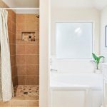 A bright bathroom with a tiled walk-in shower, a white bathtub, a white vanity with a sink and mirror, a frosted window, and a green plant on the tub ledge.