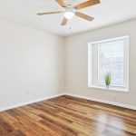 A bright, empty room with light beige walls, wood flooring, a ceiling fan with light, and a window with blinds. A small potted plant sits on the windowsill.