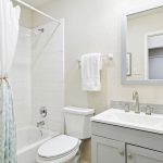 A bright, modern bathroom with a bathtub and shower, white curtain, toilet, light gray vanity with dual sinks, a large mirror, towels, and a small potted plant on the countertop.
