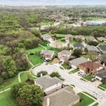 Aerial view of a suburban neighborhood with houses, green lawns, trees, and winding streets bordering a wooded area, under a cloudy sky.