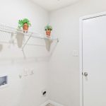 A clean, empty laundry room with a white door, a wire shelf holding four small potted plants, and hookups for a washer and dryer against a white wall.